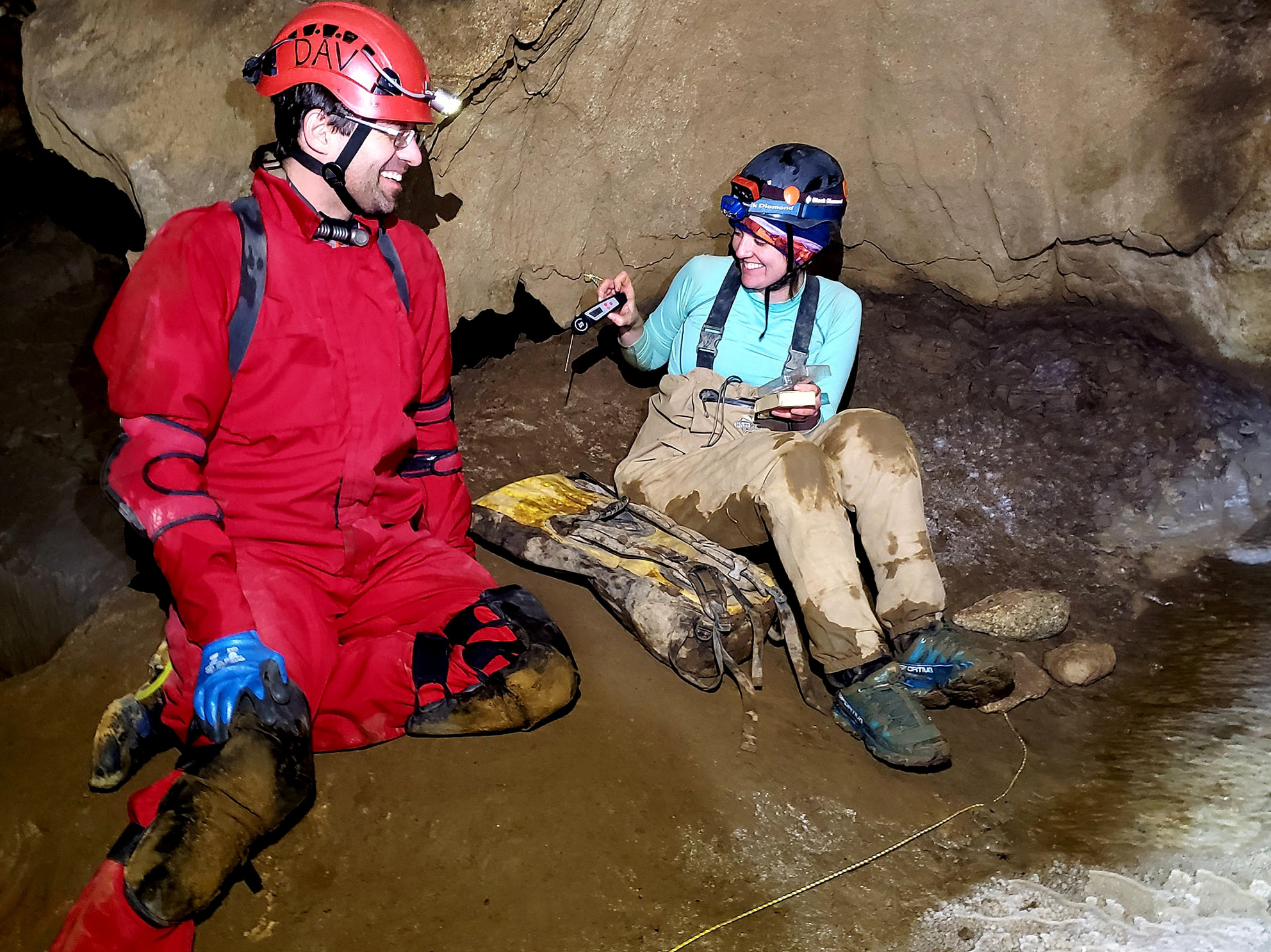 Two scientists wearing hard hats and hip waders sit in a cave