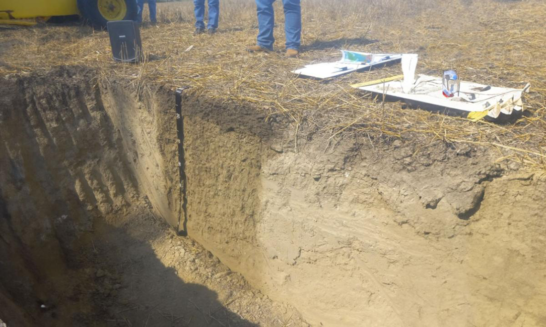 A pit with a vertical wall dug into soil to expose the layers. Instruments and leg + feet of people are on the side.