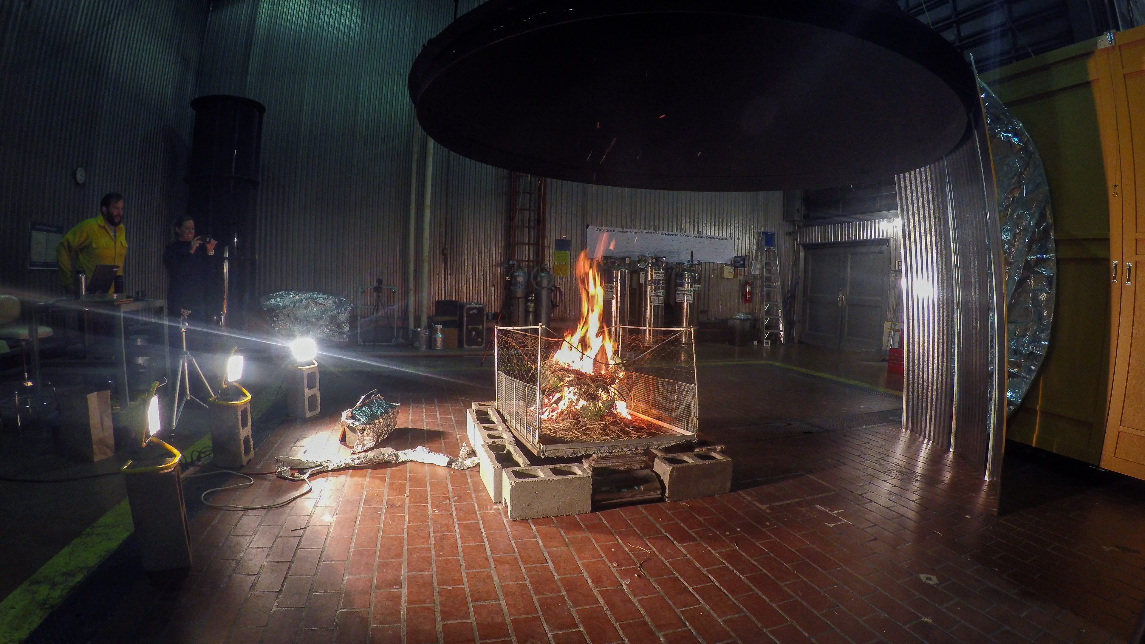 in a darkened lab, flood lights illuminate a fire burning on a platform beneath a very large, circular vent hood